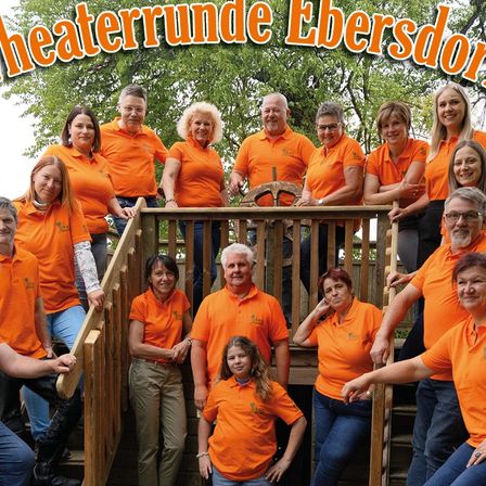 Eine Gruppe von Menschen in orangefarbenen Hemden posiert auf einer Holztreppe mit einem Hintergrund aus Bäumen.