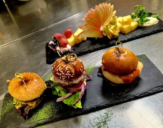 Drei Burger auf schwarzen Schieferplatten, einer mit grüner Garnierung und ein anderer mit roter Sauce, mit Obst und Käse auf einer schwarzen Schieferplatte im Hintergrund.