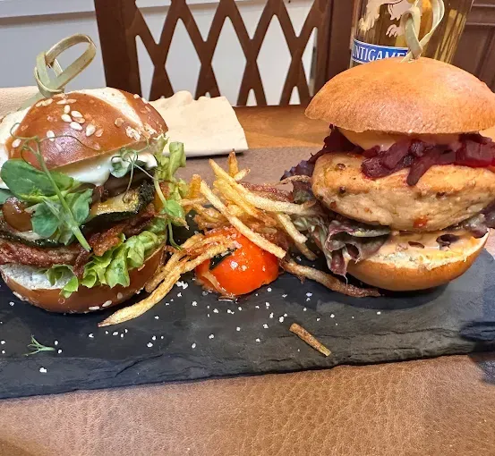 Zwei Gourmet-Burger stehen auf einem Holztisch, einer mit Gemüse und der andere mit Hühnchen. Jeder Burger wird von Pommes und einer kleinen Tomate begleitet.