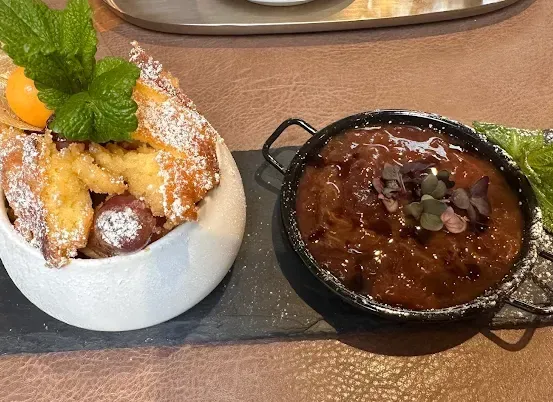 Eine Dessert-Präsentation mit einer weißen Schüssel mit Gebäck, bestreut mit Puderzucker, einer Minzblatt und einer Zitronenscheibe. Daneben steht eine schwarze Schüssel mit einer reichhaltigen, braunen Sauce, garniert mit kleinen, grünen Blättern.
