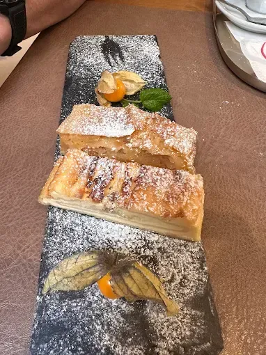 Zwei Stücke Apfelstrudel mit Puderzucker bestreut liegen auf einem schwarzen rechteckigen Teller, dekoriert mit Blättern und Früchten.
