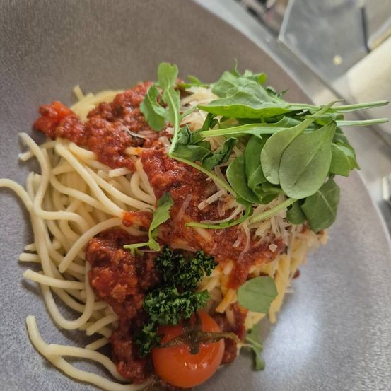 Ein Teller mit Spaghetti, überzogen mit Tomatensauce, geriebenem Käse, Rucola, Brokkoli und einer Kirschtomate.