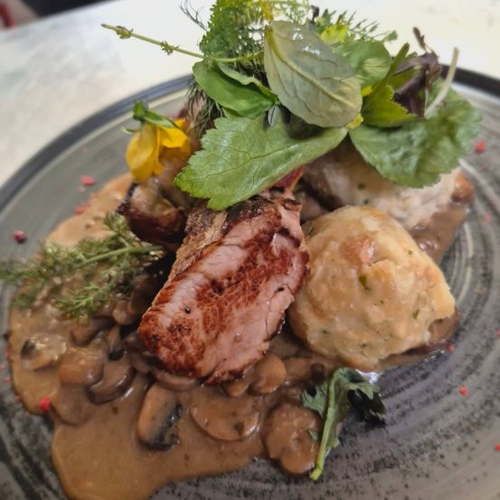 Ein Teller mit gebratenem Schweinefleisch in Pilzsauce, serviert mit Knödeln und grünen Blättern. Das Gericht ist mit einer gelben Blume garniert.
