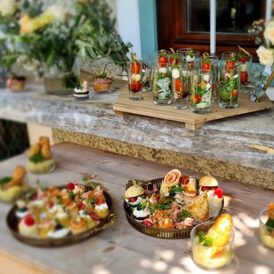 Ein Holztisch zeigt eine Auswahl an Mini-Sandwiches und Vorspeisen, darunter Räucherlachs, Käse und Gemüse, auf Tellern und in Gläsern, mit Blumen im Hintergrund.