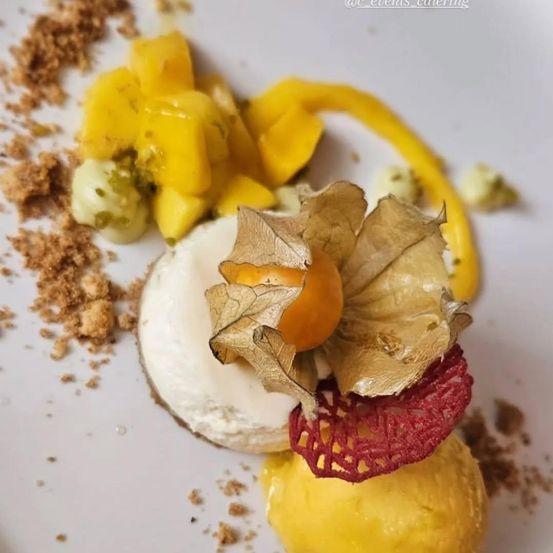 Auf einem Teller mit einem Dessert befinden sich ein gelb-orangefarbenes Fruchtpüree, ein Käseball und eine Garnitur aus goldenen Blättern. Der Teller ist mit Krümeln und braunem Pulver verziert, und eine gelbe Sauce träufelt über das Dessert.