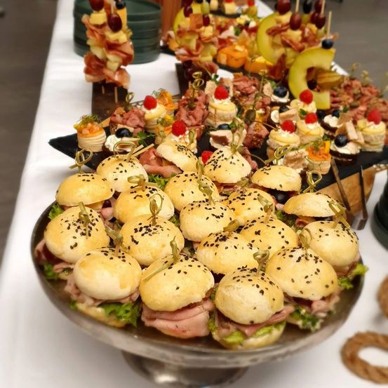 Ein Tisch mit verschiedenen Speisen, darunter Sandwiches, Fleischspieße und Desserts, auf Tellern mit schwarzen Tabletts angeordnet.