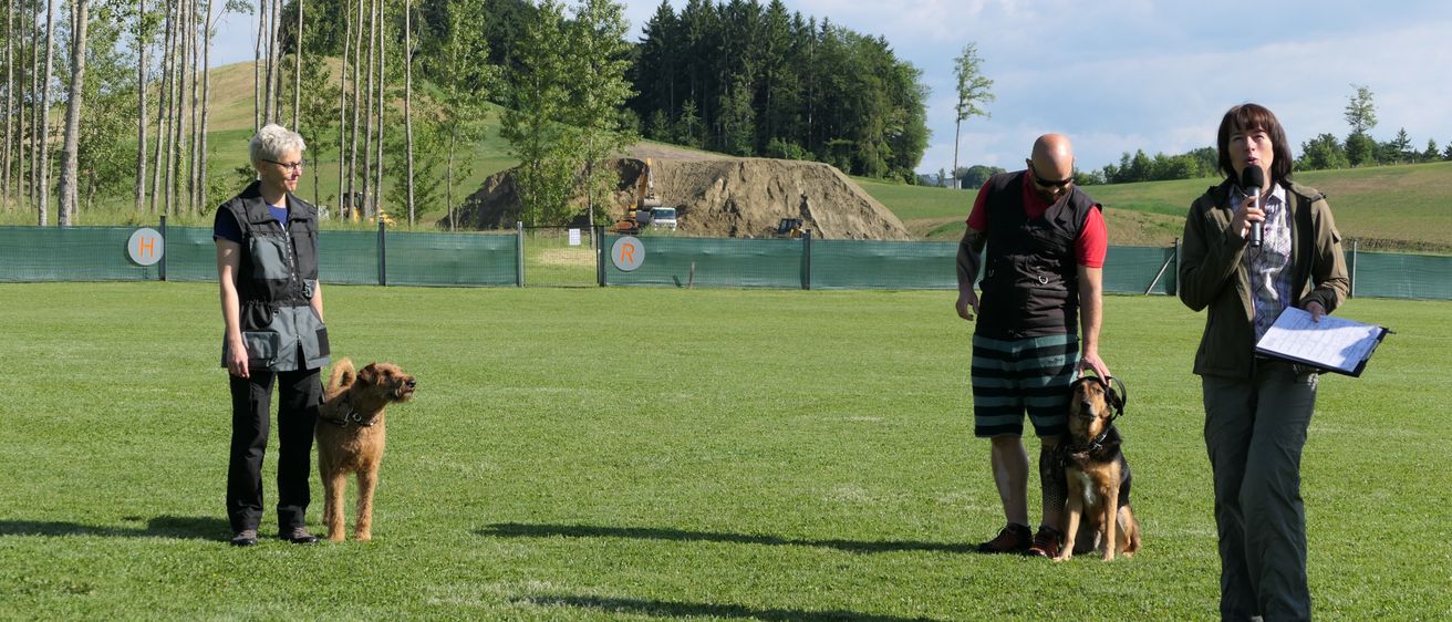 Eine Frau und ein Mann mit Hunden auf einem Feld mit einem Erdhügel im Hintergrund und einem Schild davor.