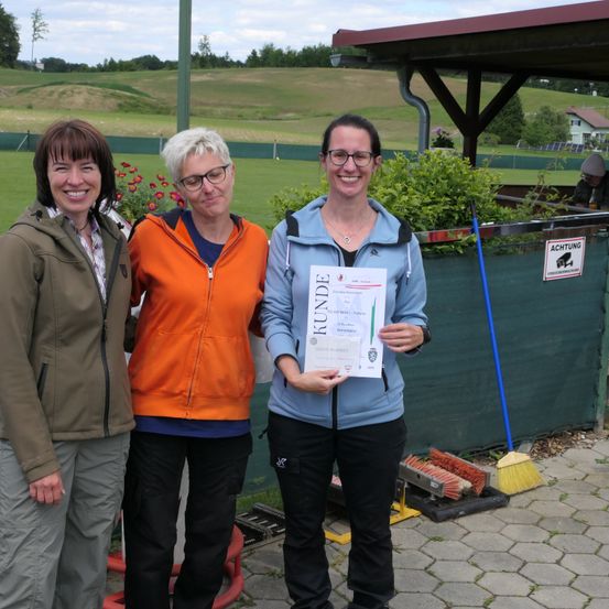 Drei Frauen stehen nebeneinander und lächeln. Die Frau in der Mitte hält ein Zertifikat mit dem Wort Kunde darauf. Sie stehen vor einem grünen Zaun.