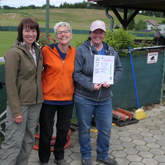 Drei Frauen stehen bei einer Veranstaltung im Freien. Eine hält ein Zertifikat. Sie lächeln und stehen vor einem grünen Zaun mit einem Schild. Ein Rasenfeld und ein Haus sind im Hintergrund.