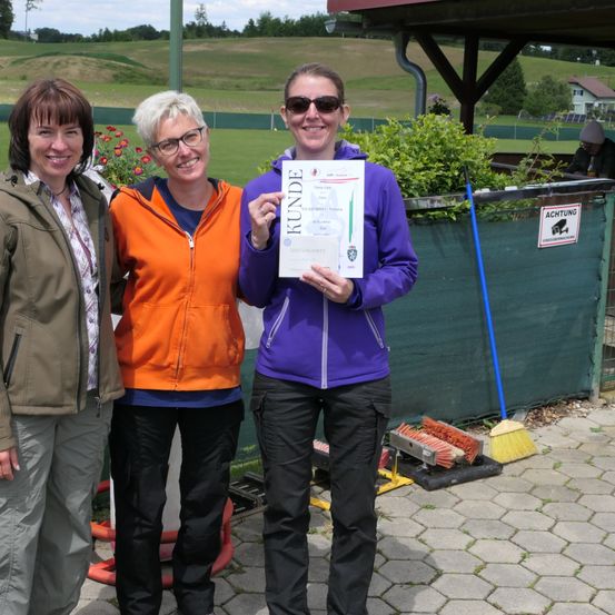 Drei Frauen stehen zusammen auf einem Weg, halten eine Urkunde, mit einer grün bedeckten Struktur im Hintergrund.