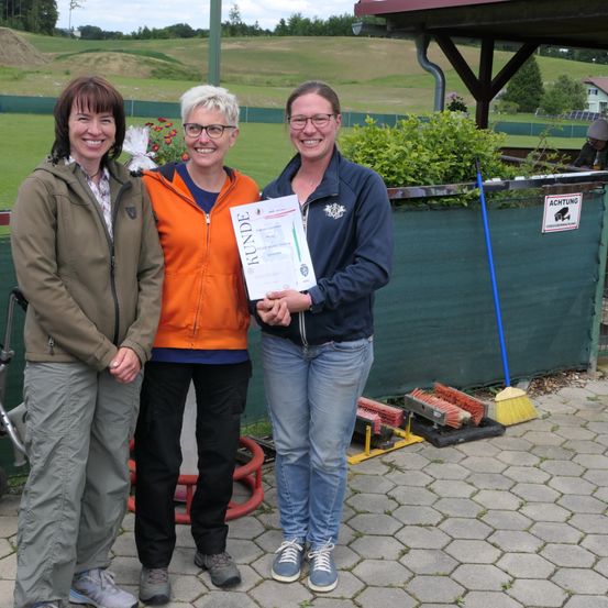 Drei Frauen stehen lächelnd vor einem grünen Zaun, eine hält ein Zertifikat. Dahinter ein Feld mit Grünpflanzen und ein Gebäude.