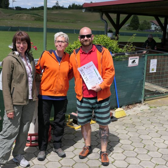 Drei Personen stehen auf einem gepflasterten Bereich in der Nähe eines Grasfeldes. Der Mann in der Mitte hält eine Urkunde. Dahinter befindet sich ein grüner Zaun mit einem Schild.