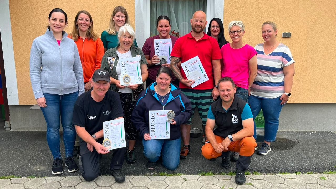 Eine Gruppe Erwachsener posiert für ein Foto, hält Urkunden und Medaillen. Einige lächeln und tragen eine Brille. Ein Mann im roten Shirt hat Tattoos am Arm.