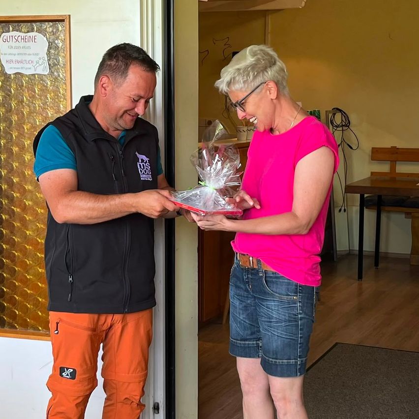 Ein Mann mit schwarzem Weste und orangefarbener Hose übergibt ein Geschenk an eine Frau in einem rosa T-Shirt. Sie stehen vor einer Tür, mit einem Schild und einer Bank dahinter.