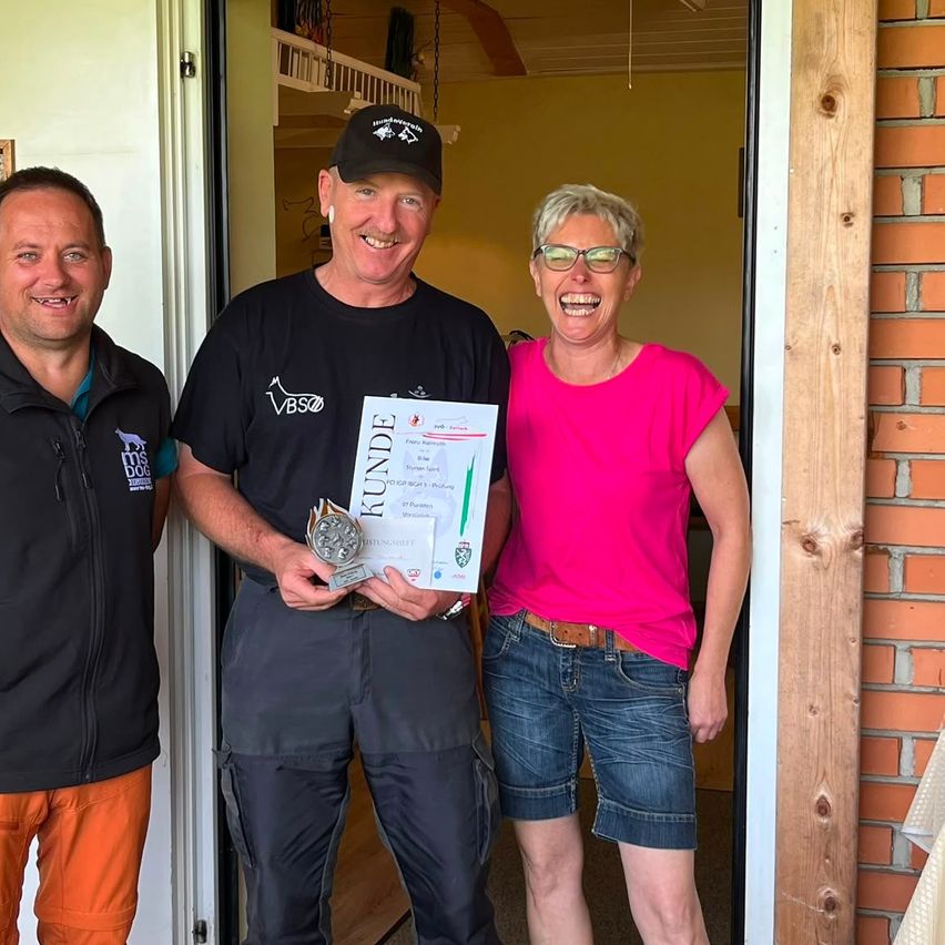 Drei Personen stehen lächelnd in einer Tür. Der Mann in der Mitte hält einen Pokal und ein Zertifikat. Eine Frau in einem rosa T-Shirt und ein Mann in einem schwarzen Jackett begleiten ihn.