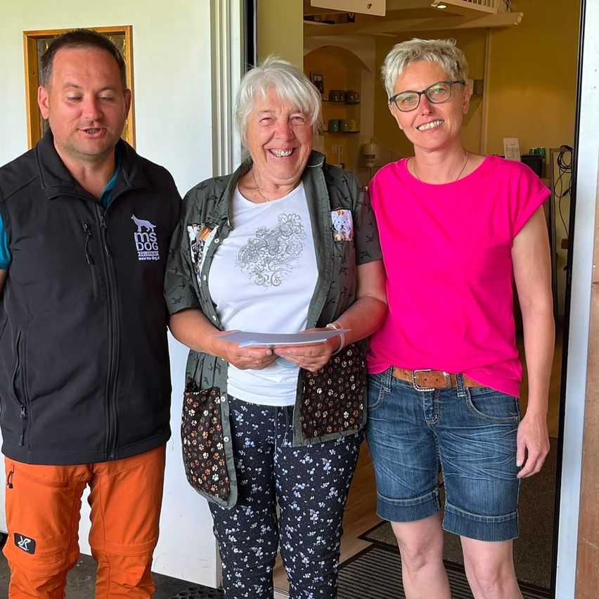 Drei Menschen stehen zusammen und lächeln, einer hält Papiere. Der Mann trägt eine schwarze Jacke mit Logo, die Frau in Weiß hält Papiere und die andere Frau trägt ein rosa Shirt und eine Brille.