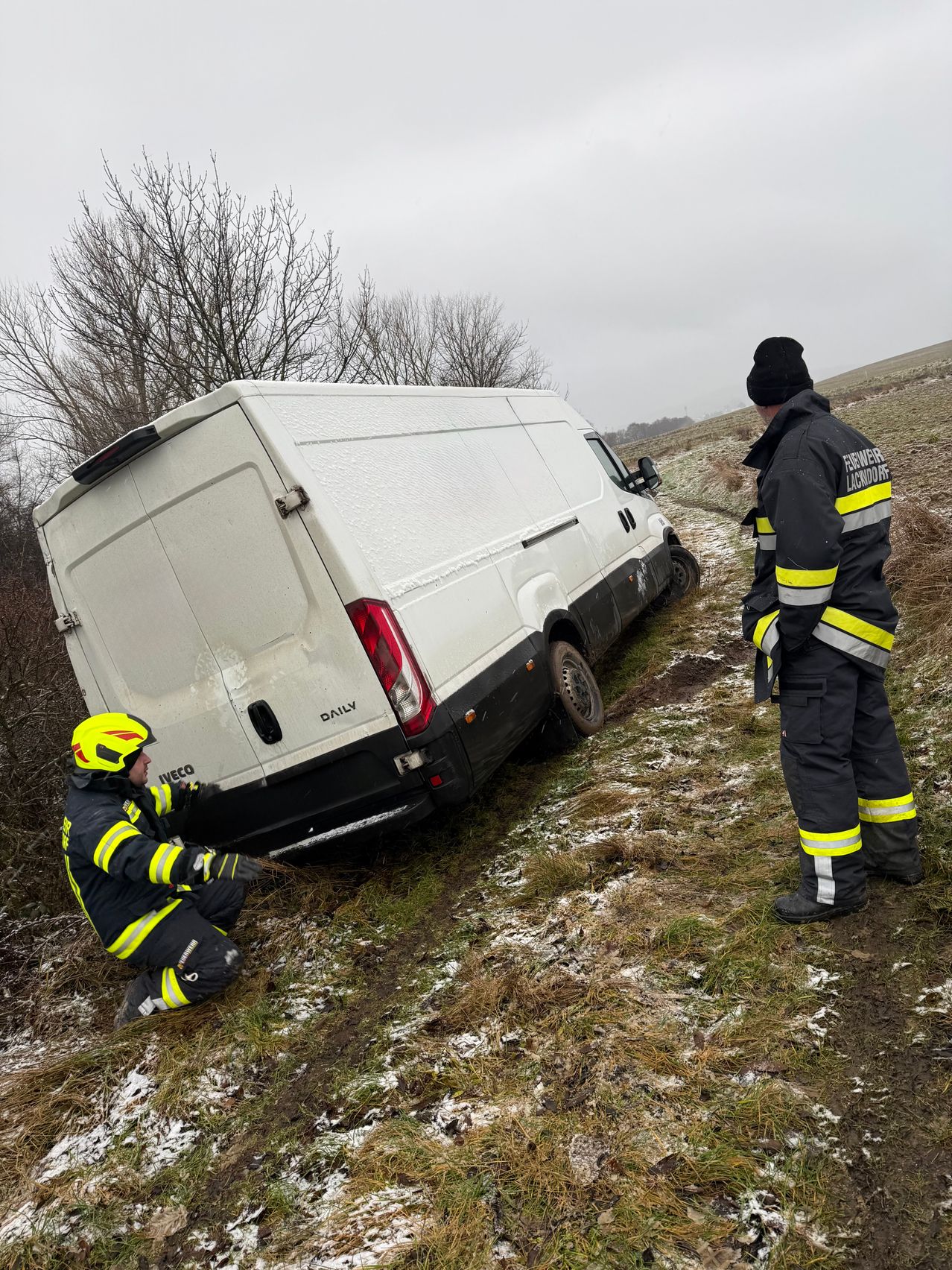 Feuerwehrleute in Schutzkleidung helfen einem festsitzenden Van auf einem verschneiten Feld, einer kniet, um von hinten zu schieben.