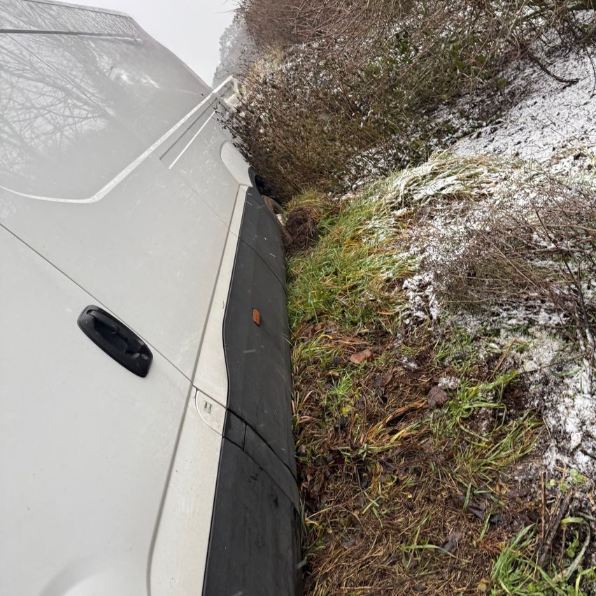 Ein weißer Lieferwagen steckt in einem Graben fest, teilweise von Schnee und Vegetation bedeckt. Die Seite des Fahrzeugs zeigt einen schwarzen Stoßfänger mit Beschädigungen.
