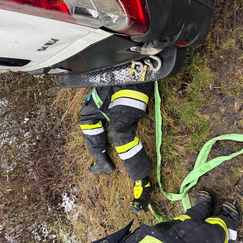 Ein Feuerwehrmann in Schutzkleidung liegt auf dem Boden mit einem Autostoßfänger darüber, verbunden durch Seile. Die Szene spielt sich im Freien auf einem Grasfeld ab.