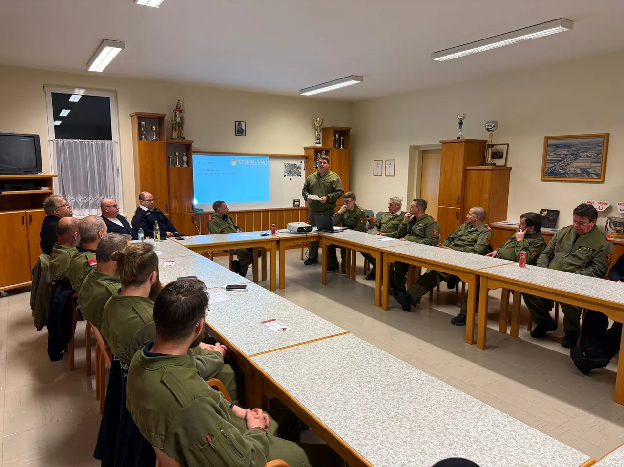 Eine Gruppe von Menschen in Uniform sitzt um einen Tisch in einem Raum mit Beamerleinwand und einem stehenden Mann.