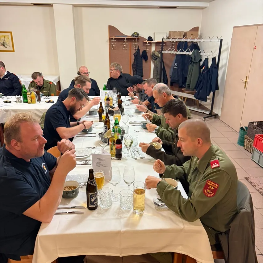 Eine Gruppe von Männern, einige in Uniform, sitzen um einen langen Tisch und essen und trinken. Flaschen mit Bier und Weingläser stehen auf dem Tisch. Der Raum hat geflieste Böden und eine Holztür.