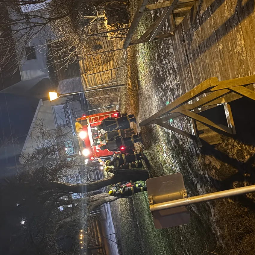 Feuerwehrleute in Schutzkleidung stehen neben einem roten Feuerwehrwagen mit blinkenden Lichtern auf einer Straße bei Nacht.