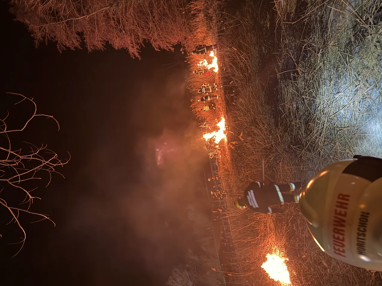 Feuerwehrleute bekämpfen in der Nacht ein Feuer, mit mehreren Flammen in einem Feld. Ein Feuerwehrmann steht in der Nähe und beobachtet die Situation.