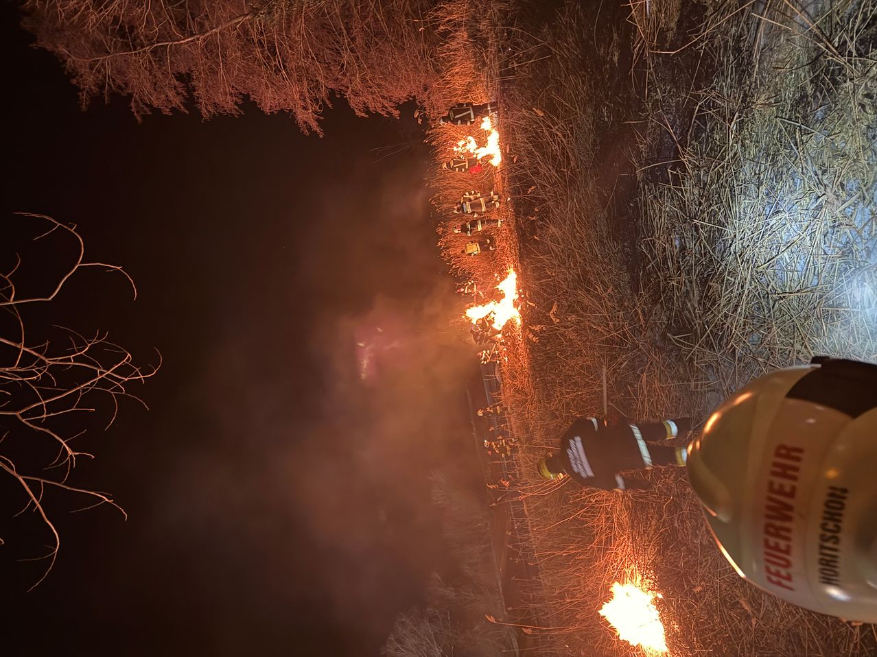Firefighters are battling a blaze at night, with multiple flames in a field. A firefighter stands nearby, observing the situation.