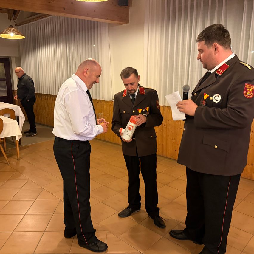 Drei Männer stehen drinnen; einer hält ein Glas, ein anderer einen Pokal und der dritte liest von einem Papier. Sie befinden sich in einem Saal mit Holzwänden und gefliestem Boden.