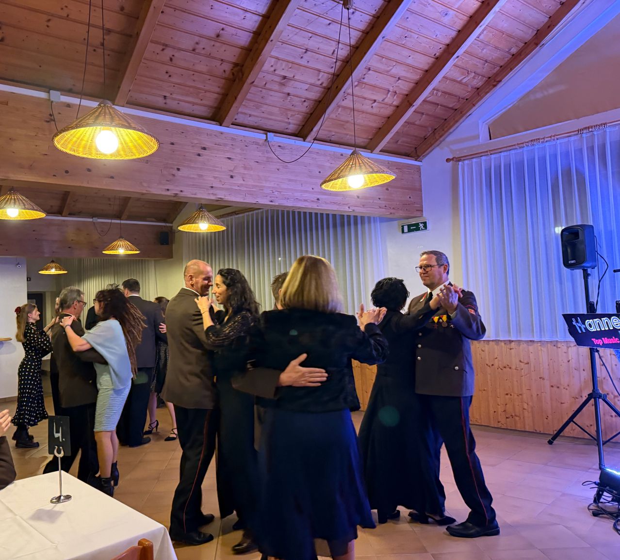 In einem Bankettsaal tanzen mehrere Paare. Der Raum hat Holzbalken und Vorhänge. Ein Lautsprecher steht rechts.