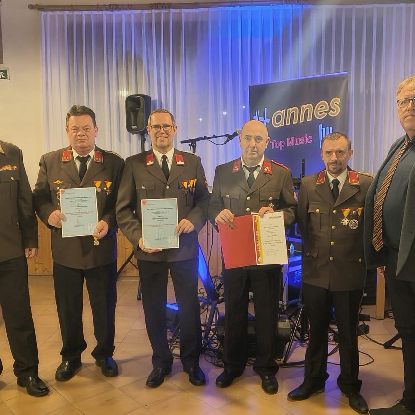 Fünf Männer in Militäruniformen stehen zusammen und halten Urkunden. Dahinter befindet sich eine Bühne mit Mikrofon und Banner.