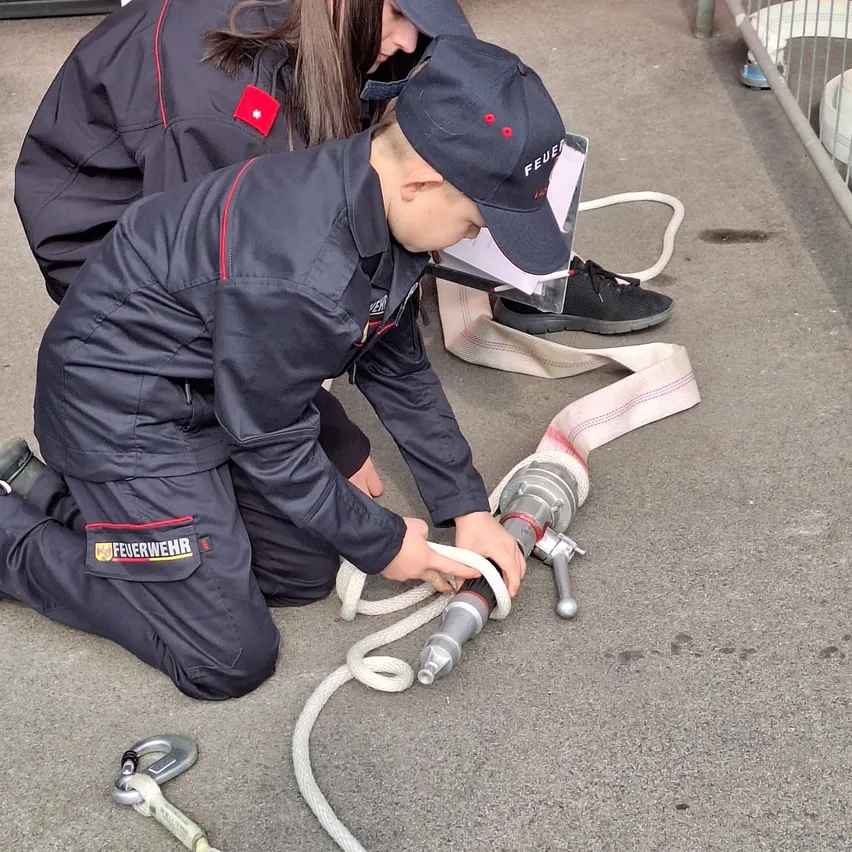 Ein junger Junge in Feuerwehruniform kniet auf dem Boden und justiert einen Feuerwehrschlauch. Eine Frau in Uniform beobachtet ihn, beide tragen Schutzausrüstung.