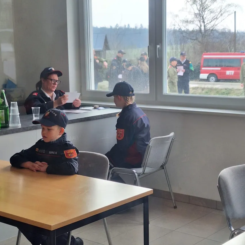 Eine Gruppe von Personen in Uniformen, einschließlich einer Frau und eines jungen Jungen, sitzen an einem Tisch in einem Raum mit Fenstern. Draußen ist ein roter Feuerwehrwagen auf der Straße geparkt.