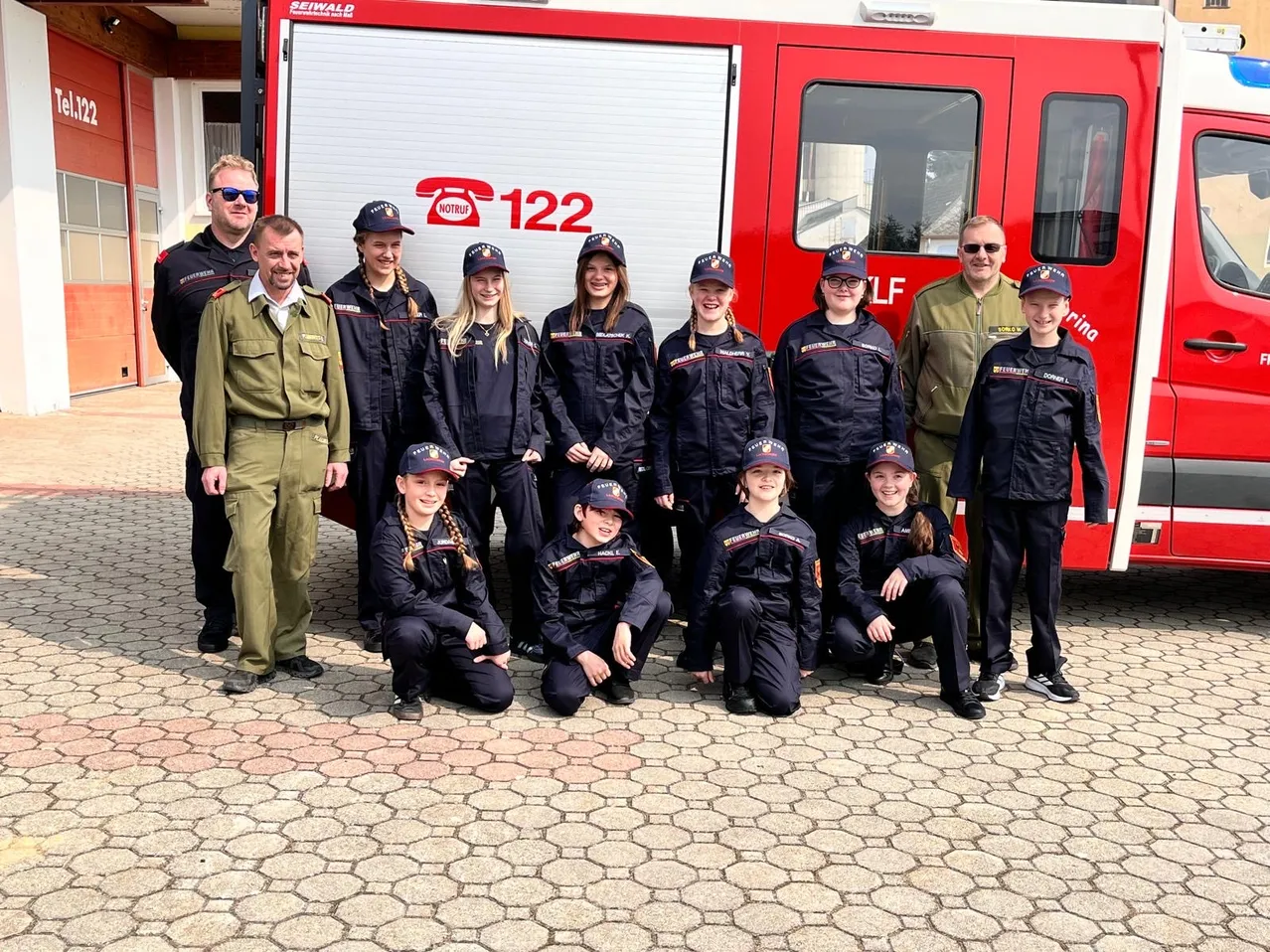 Eine Gruppe von Menschen in Uniformen posiert vor einem roten Feuerwehrauto. Es gibt zwei Männer und neun Frauen, die alle in die Kamera schauen.