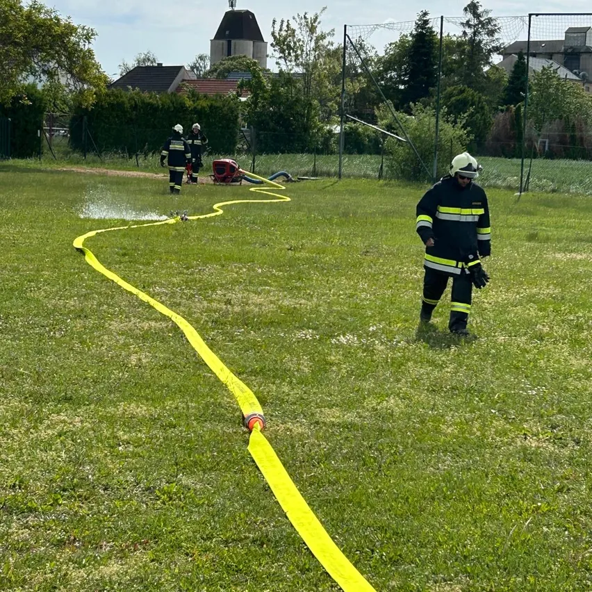 Feuerwehrleute sind auf einem Rasenfeld und halten einen gelben Schlauch. Ein Feuerwehrmann sprüht Wasser, während zwei andere in der Nähe sind. Ein Feuerwehrwagen ist geparkt. Bäume und Häuser sind im Hintergrund.
