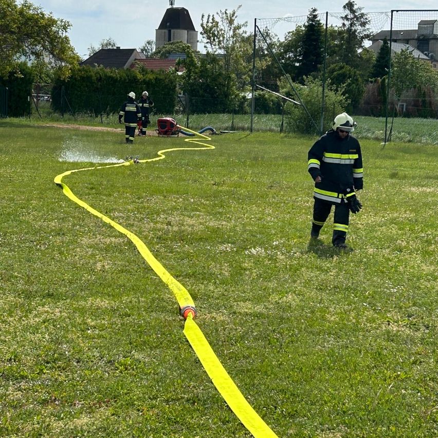 Feuerwehrleute sind auf einem Rasenfeld und halten einen gelben Schlauch. Ein Feuerwehrmann sprüht Wasser, während zwei andere in der Nähe sind. Ein Feuerwehrwagen ist geparkt. Bäume und Häuser sind im Hintergrund.