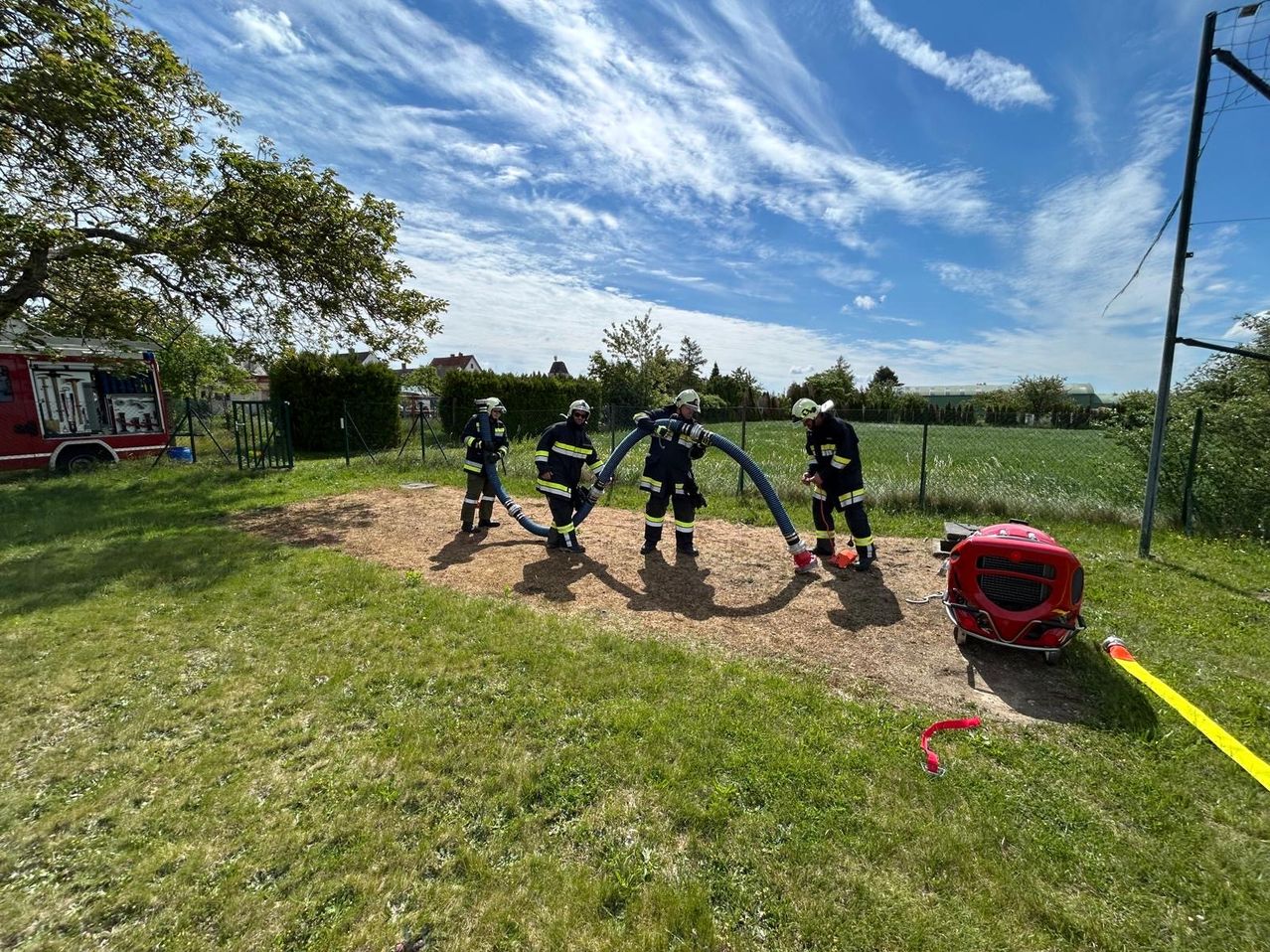 Vier Feuerwehrleute in voller Ausrüstung stehen in einem Feld mit Schläuchen, die an einem Hydranten angeschlossen sind. Eine rote Ausrüstung ist in der Nähe geparkt. Bäume und ein Zaun sind im Hintergrund.