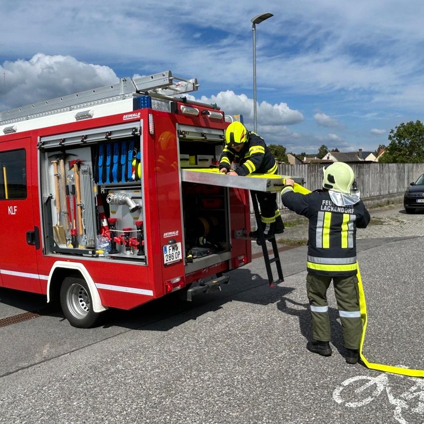 Zwei Feuerwehrleute arbeiten an einem roten Feuerwehrauto. Einer steht auf einer Leiter und der andere klettert hoch. Das Auto ist auf der Straße geparkt.