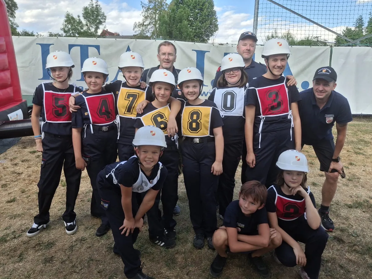 Eine Gruppe junger Kinder in Sportuniformen und Helmen, nummeriert von 0 bis 9, posiert für ein Foto mit zwei Erwachsenen vor einem Banner.