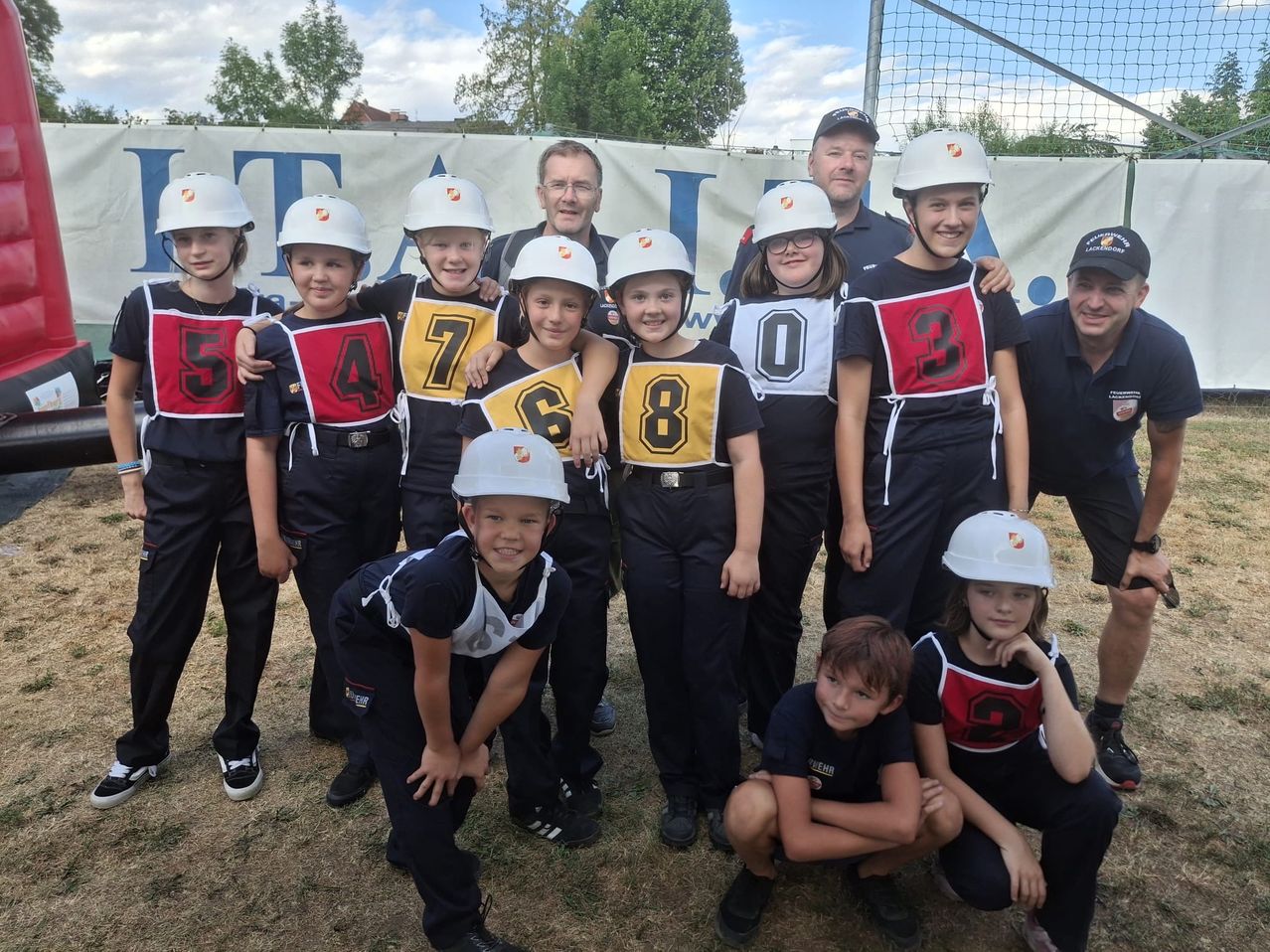Eine Gruppe junger Kinder in Sportuniformen und Helmen, nummeriert von 0 bis 9, posiert für ein Foto mit zwei Erwachsenen vor einem Banner.