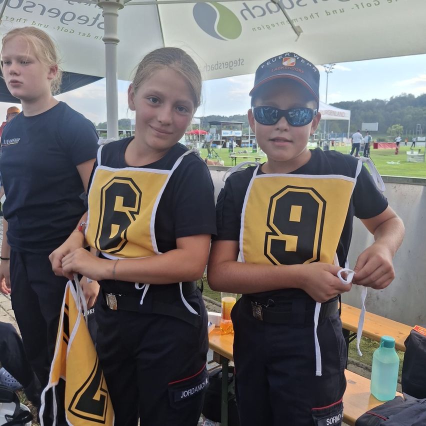 Zwei junge Kinder, ein Mädchen mit der Nummer 4 und ein Junge mit der Nummer 9 auf ihren Trikots, stehen zusammen auf einem Sportplatz. Sie tragen passende Uniformen und scheinen für ein Foto zu posieren.