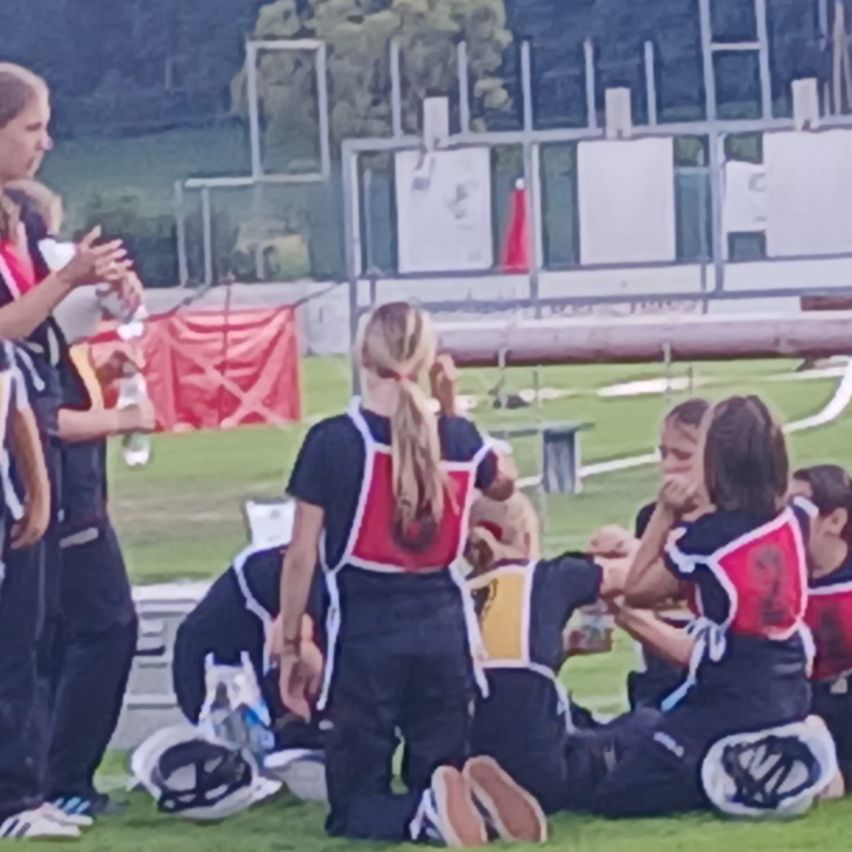 Eine Gruppe junger Athleten in Uniform befindet sich auf einem Rasenfeld, einige sitzen und andere stehen, mit einem verschwommenen Hintergrund aus Metallstrukturen und Bäumen.