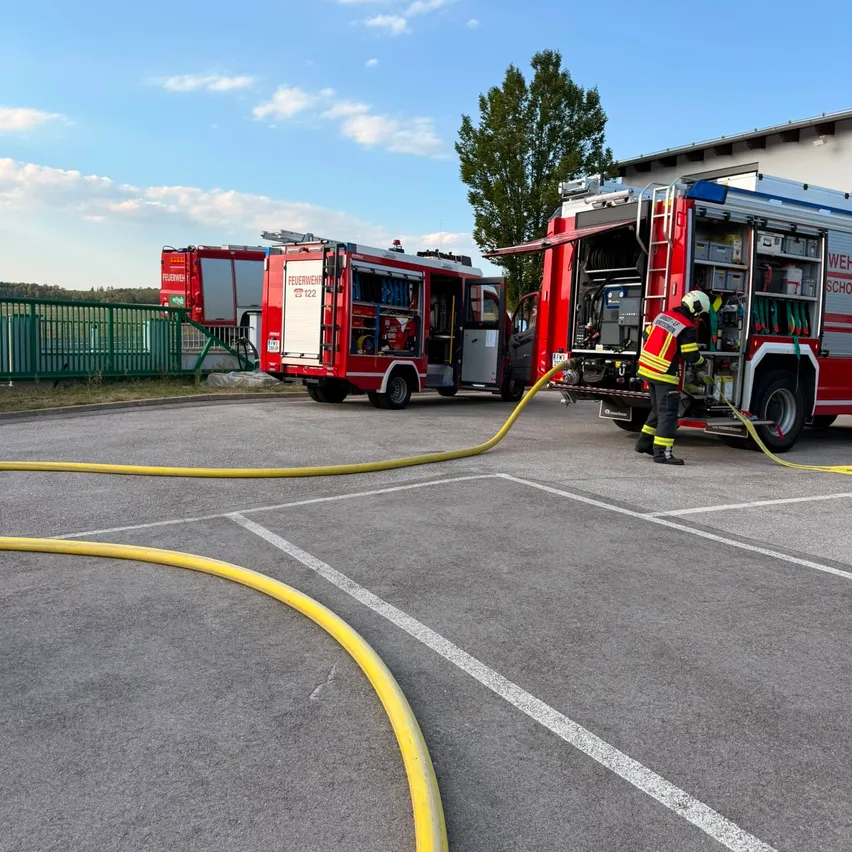 Ein Feuerwehrmann verbindet einen Schlauch mit einem Feuerwehrauto auf einem Parkplatz, wobei zwei weitere Trucks im Hintergrund stehen.