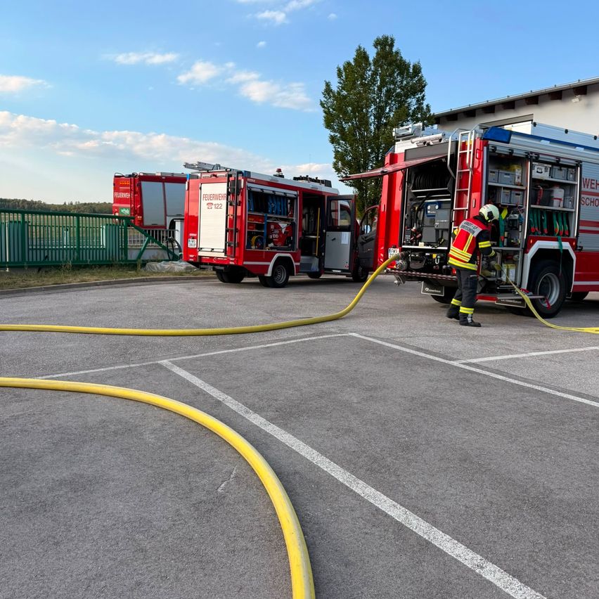Ein Feuerwehrmann verbindet einen Schlauch mit einem Feuerwehrauto auf einem Parkplatz, wobei zwei weitere Trucks im Hintergrund stehen.