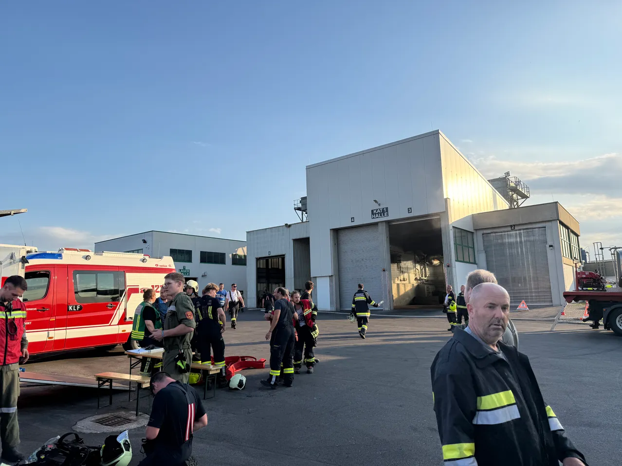 Eine Gruppe von Feuerwehrleuten in Uniform versammelt sich vor einem großen Gebäude mit offener Garagentür. Ein roter und weißer Krankenwagen ist in der Nähe geparkt.
