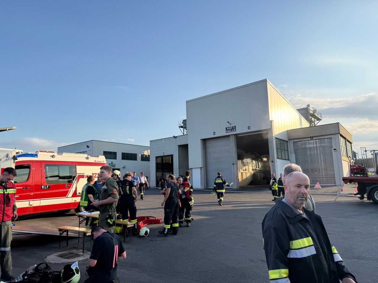 Eine Gruppe von Feuerwehrleuten in Uniform versammelt sich vor einem großen Gebäude mit offener Garagentür. Ein roter und weißer Krankenwagen ist in der Nähe geparkt.