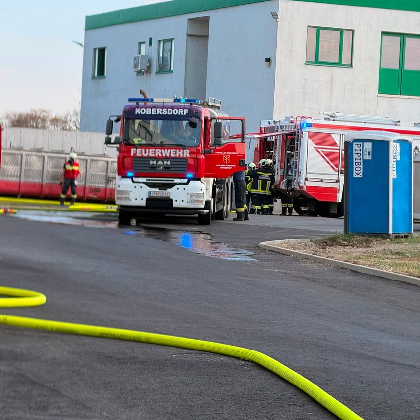 Ein Feuerwehrauto mit dem Wort 'Feuerwehr' steht in der Nähe eines Gebäudes, während Feuerwehrleute sich darauf vorbereiten, einen anderen LKW zu betreten. Ein gelber Schlauch liegt auf der nassen Straße.