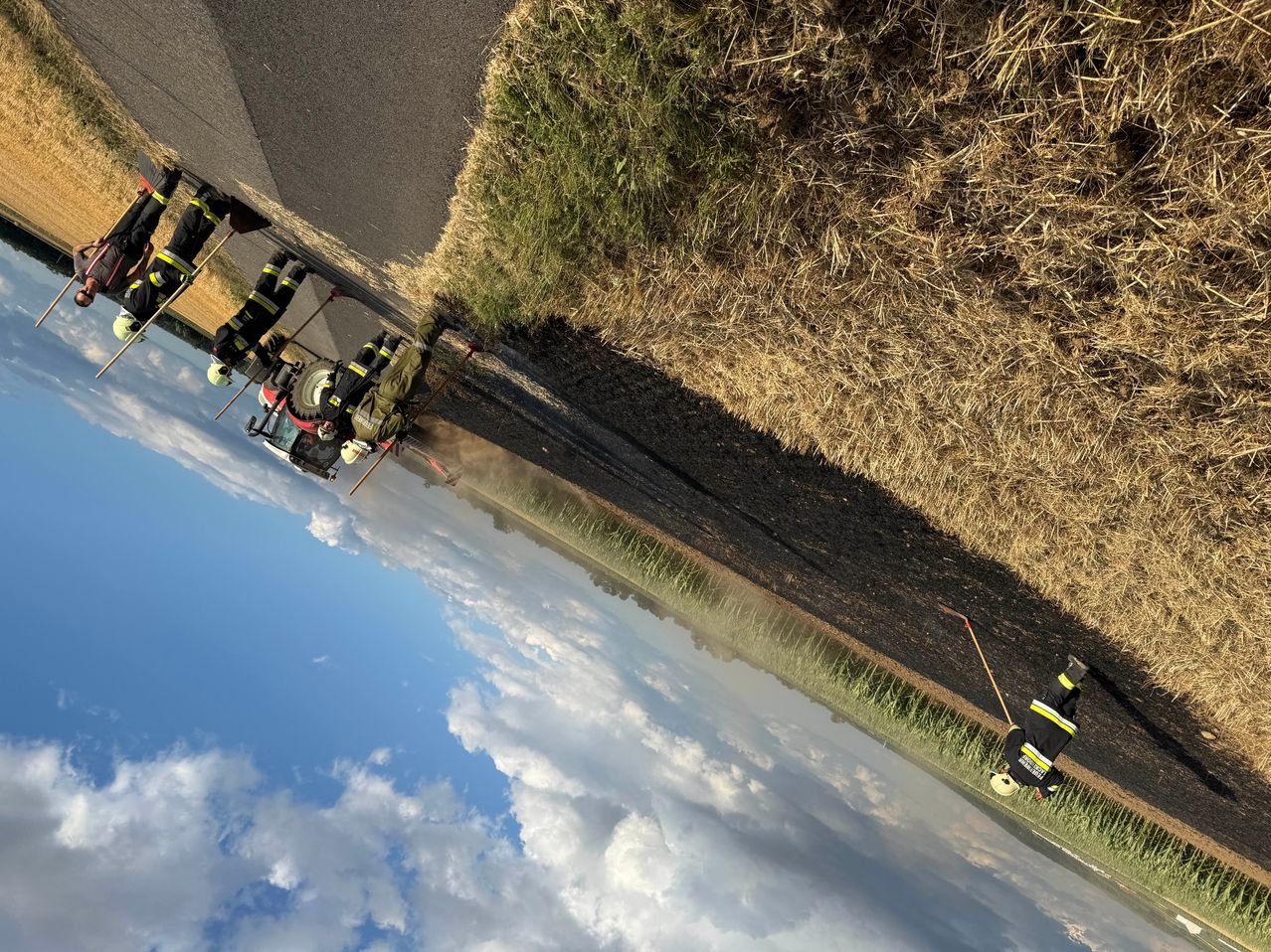 An aerial view shows firefighters wearing helmets and uniforms, clearing debris on the side of a road with a clear blue sky and clouds in the background.