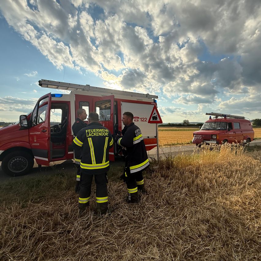 Three firefighters stand by a fire truck. The truck has the number 122 on the side. Another fire truck is parked behind them. The sky is cloudy.