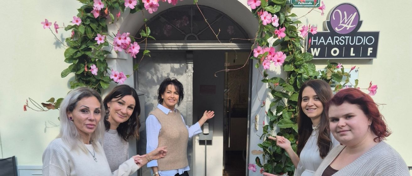 Fünf Frauen posieren vor einem weißen Gebäude, das mit rosa Blumen und einem Schild mit der Aufschrift 'Haarstudio Wolff' geschmückt ist. Die Frau in der Mitte gestikuliert, während die anderen lächeln.